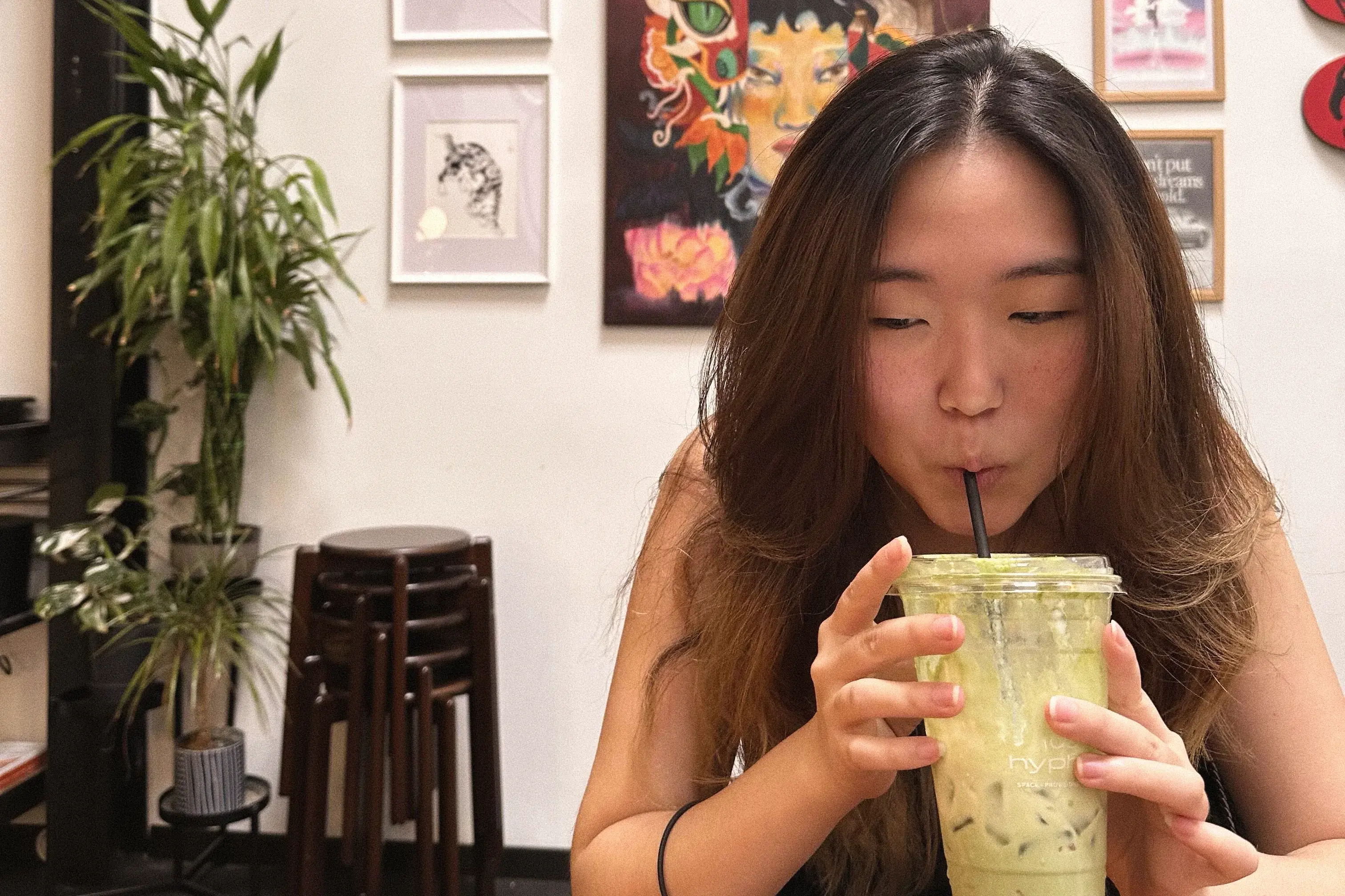 Picture of Sumin drinking an Iced matcha drink from her cup. There are paintings in the background.